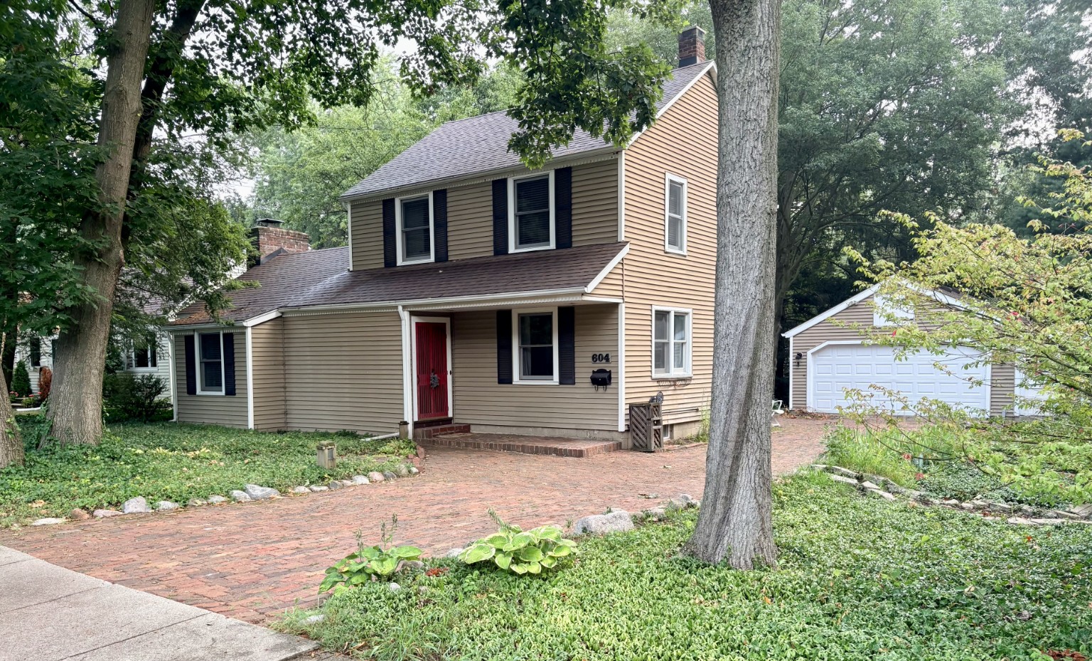 604 Jenks Boulevard from the street: a two-story colonial with dark shutters and a red front door, set back behind mature hardwoods, with a reclaimed brick paver drive leading to a detached garage.